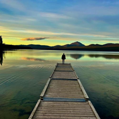 A person standing on the end of a long dock overlooks a large lake