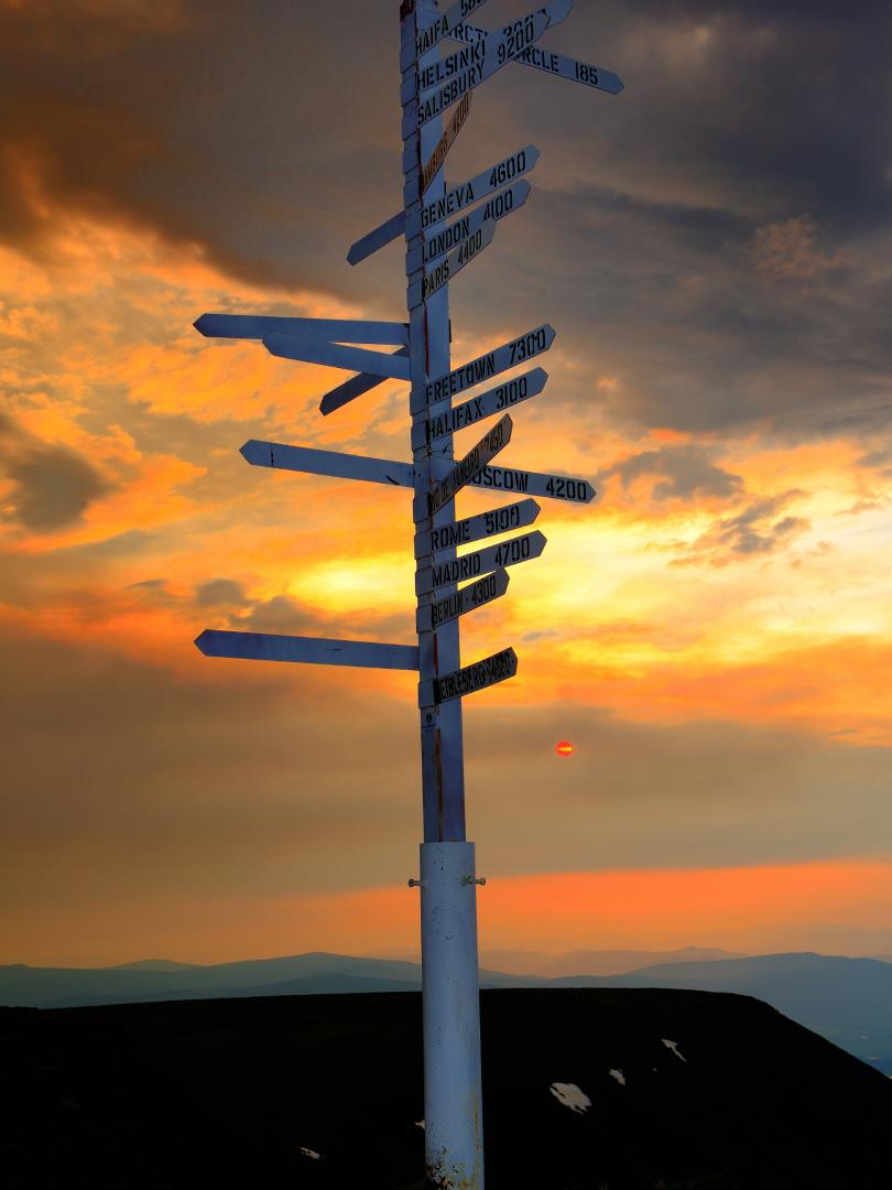 A sign post pointing to cities in many directions stands on a mountaintop at sunset