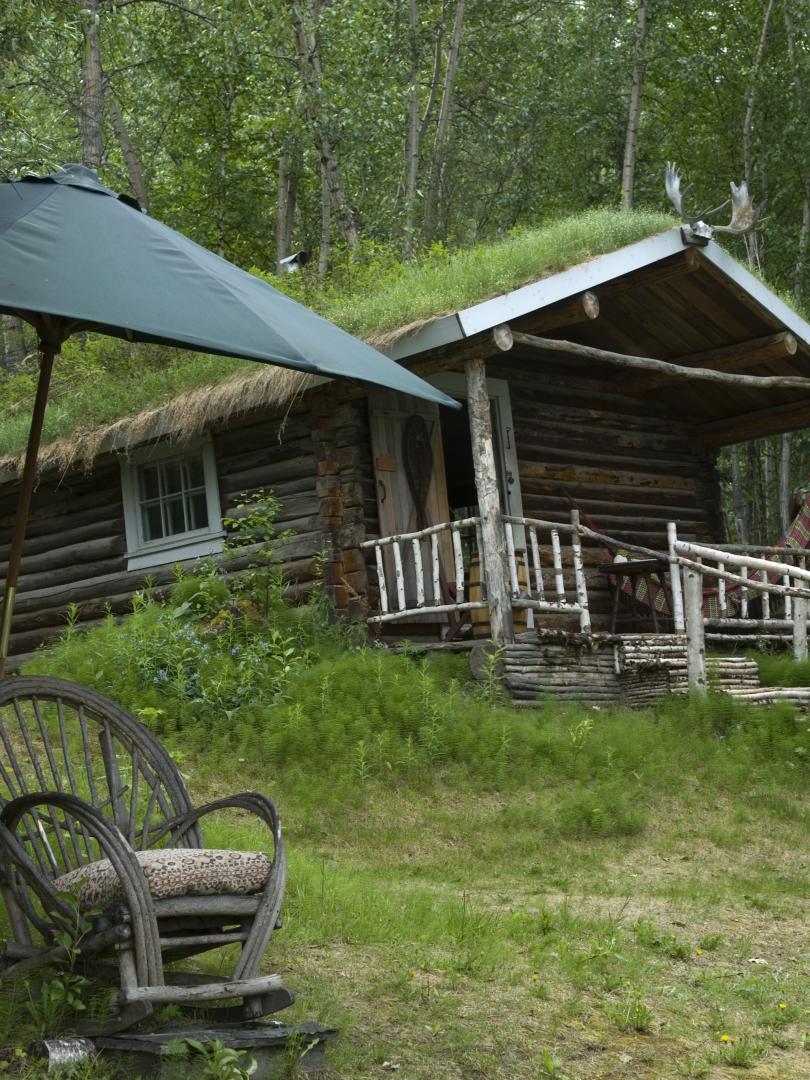 A small cabin sits on a green hillside behind a wicker chair and a large umbrella