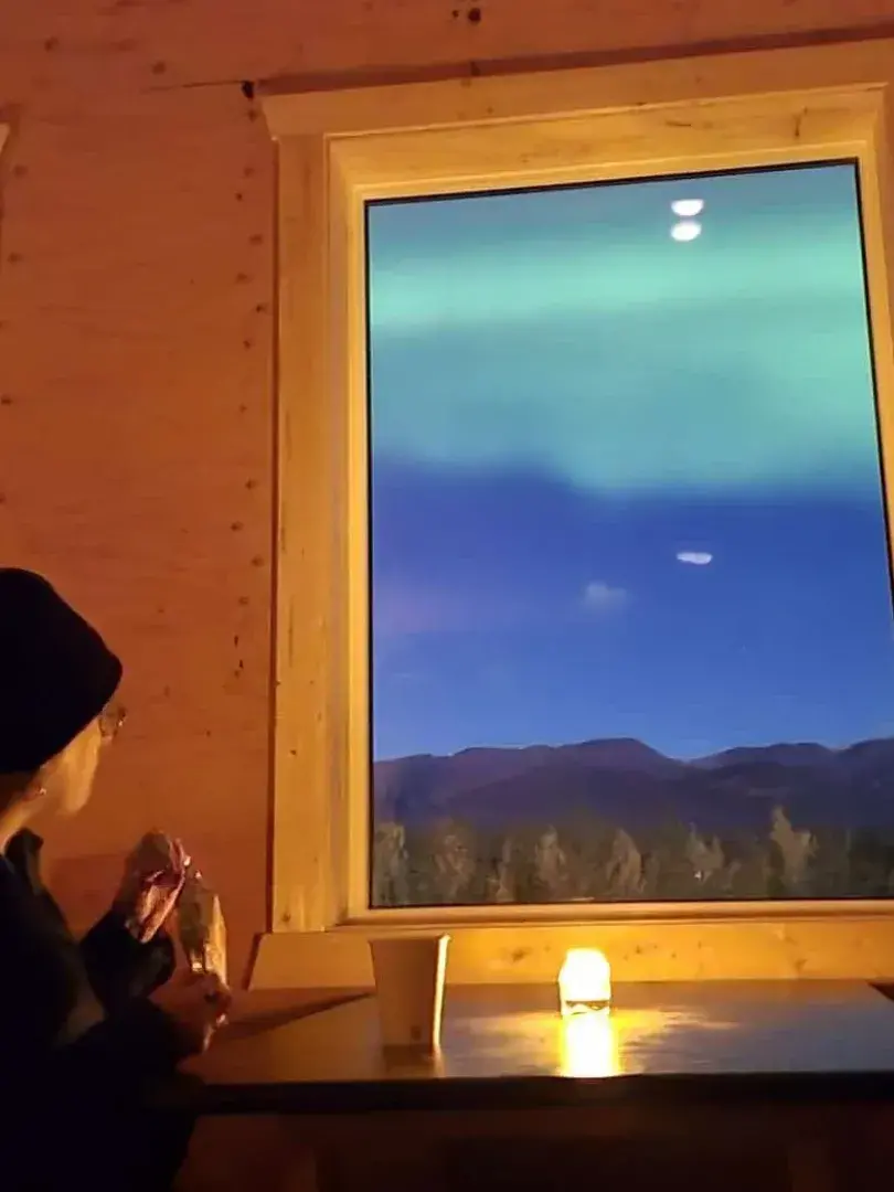 A lady sits at a table in a warm cabin, enjoying the aurora