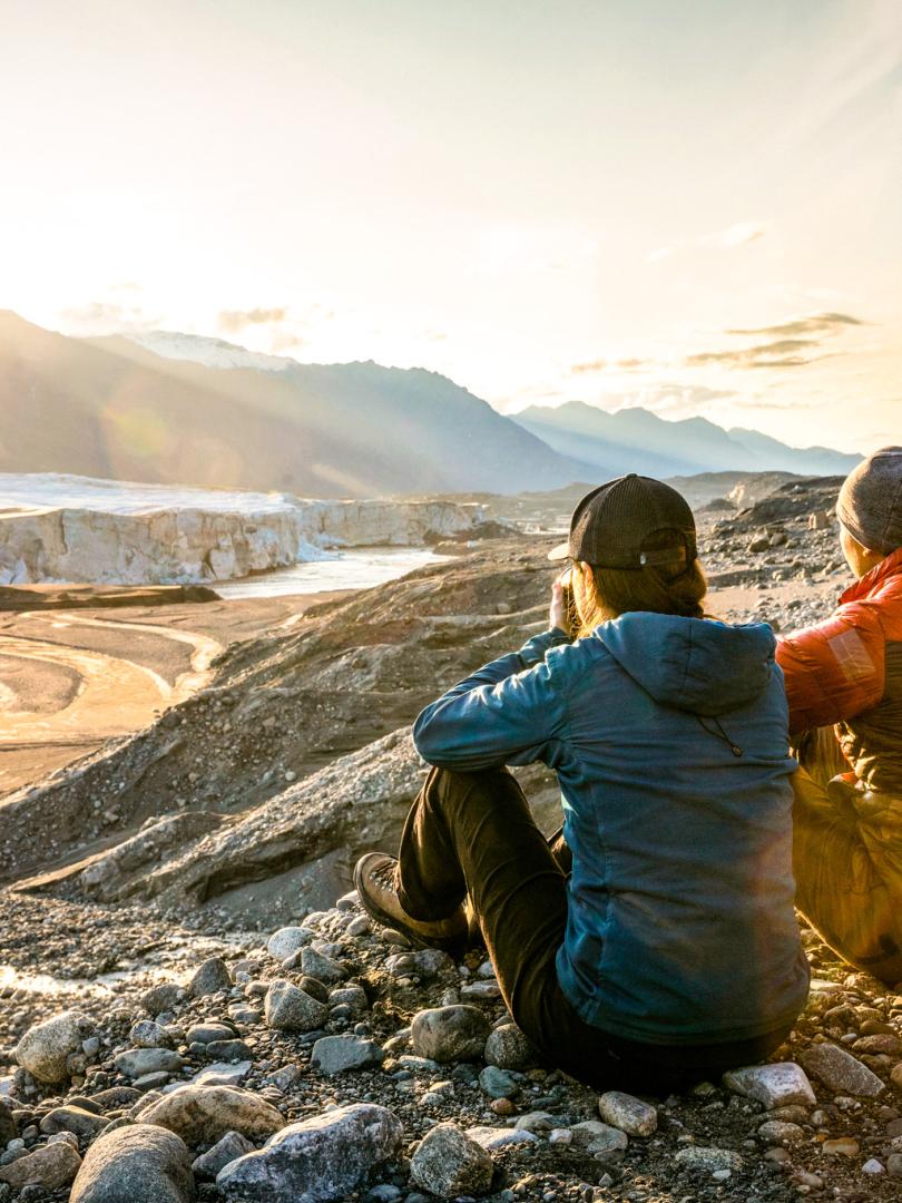 Two hikers stand in awe, gazing at the breathtaking Donjek Glacier in the distance. Surrounded by towering mountains, the glacier stretches out before them, its icy expanse shimmering under the soft light. The hikers, dressed in outdoor gear, take in the serene yet powerful beauty of the landscape, creating a sense of tranquility and connection with nature.