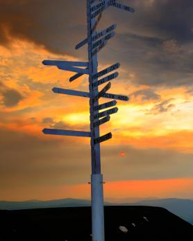 A sign post pointing to cities in many directions stands on a mountaintop at sunset