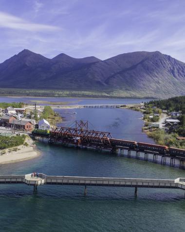 A bridge and railroad crossing extend over a lake with a small town to the left and mountains to the back