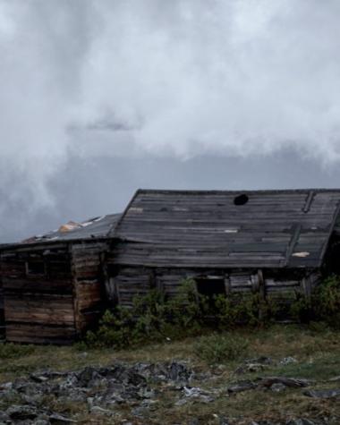 A grey cabin sits atop a mountain under low cloud coverage.